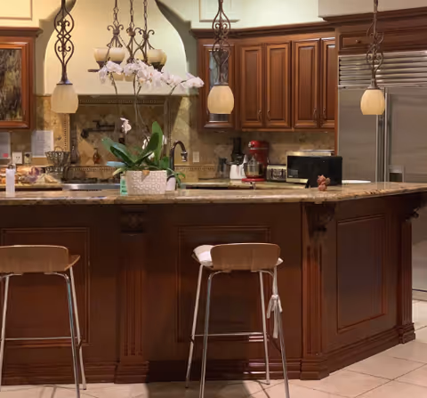 A kitchen interior featuring a large island with a granite countertop and two wooden bar stools. The island has a white orchid plant in a decorative pot. Above the island, there are three hanging pendant lights with ornate metal fixtures. The kitchen has wooden cabinets, a stainless steel refrigerator, a microwave, and various small appliances including a red stand mixer. The floor is tiled and the overall color scheme is warm with brown and beige tones.