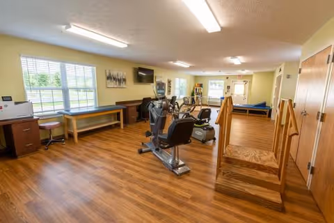 A spacious rehabilitation room with wooden flooring and light yellow walls. The room contains exercise equipment including stationary bikes and parallel bars for physical therapy. There are large windows letting in natural light, a television mounted on the wall, and various pieces of furniture such as desks and chairs. The room appears clean and well-organized.