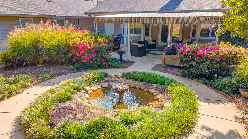 Outdoor courtyard featuring a small circular stone fountain, surrounding grasses and flowering shrubs, paved walkways, and a covered patio with seating.