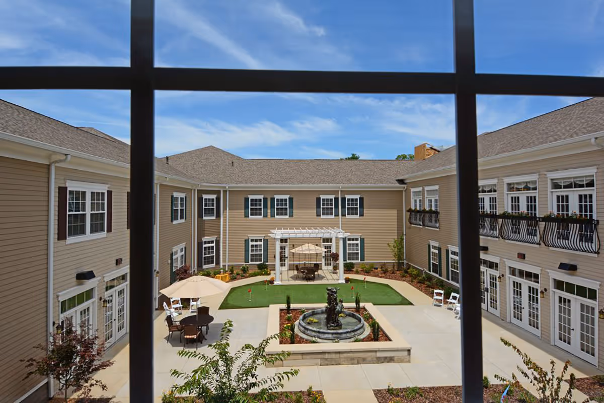 View through a window with black grid bars of a courtyard in a senior living facility. The courtyard features a central water fountain surrounded by a small garden, a putting green, patio tables with umbrellas, and beige two-story buildings with white-framed windows and doors surrounding the area under a blue sky with light clouds.