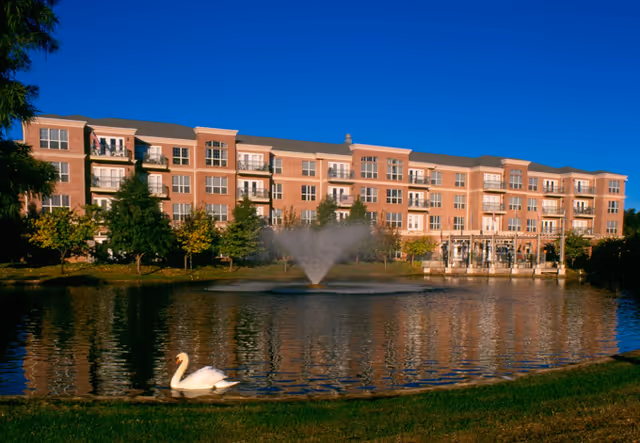 A large multi-story brick residential building with balconies overlooking a pond with a water fountain and a swan swimming near the grassy shore under a clear blue sky.