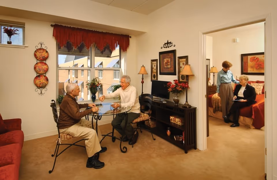 Two elderly men sitting and playing a game at a glass table in a warmly decorated living room area with a window showing an exterior building. In an adjacent bedroom, two elderly women are engaged in conversation, one standing and the other sitting on the bed. The rooms have beige walls and carpet, with decorative lamps, framed artwork, and floral arrangements.