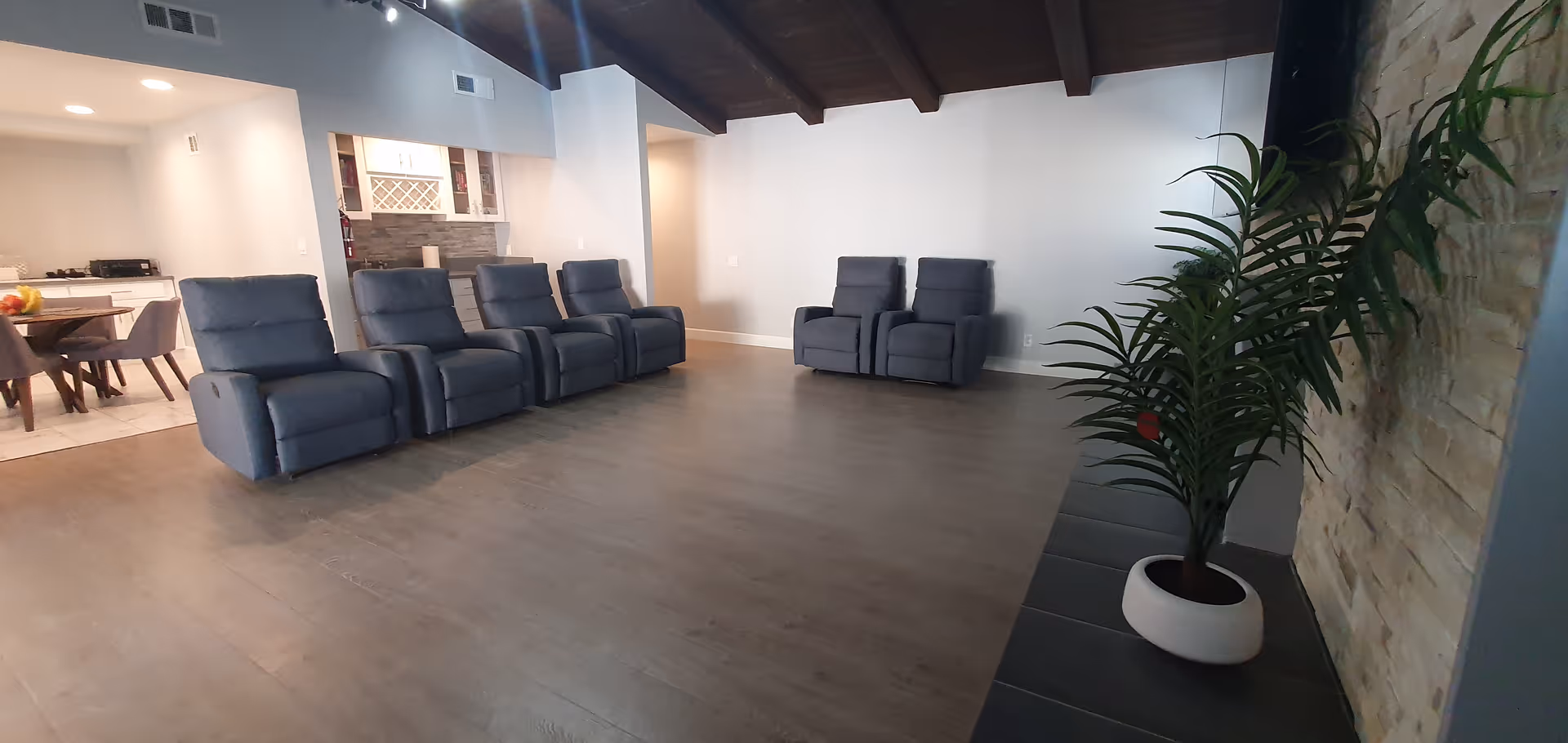 A spacious living room area with six blue recliner chairs arranged in two groups against white walls. The room has a wooden floor and a high ceiling with exposed dark wooden beams. To the left, there is a dining area with a table and chairs, and a kitchen area with white cabinets and a countertop. On the right side, there is a stone accent wall with a large potted plant in front of it.