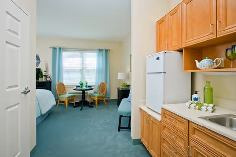 Interior view of a senior living facility room at Lorien Taneytown showing a small kitchenette area with wooden cabinets, a white refrigerator, a sink, and countertop on the right. In the background, there is a seating area with a round table and two chairs near a window with light blue curtains. Part of a bed and a nightstand with a lamp are also visible.