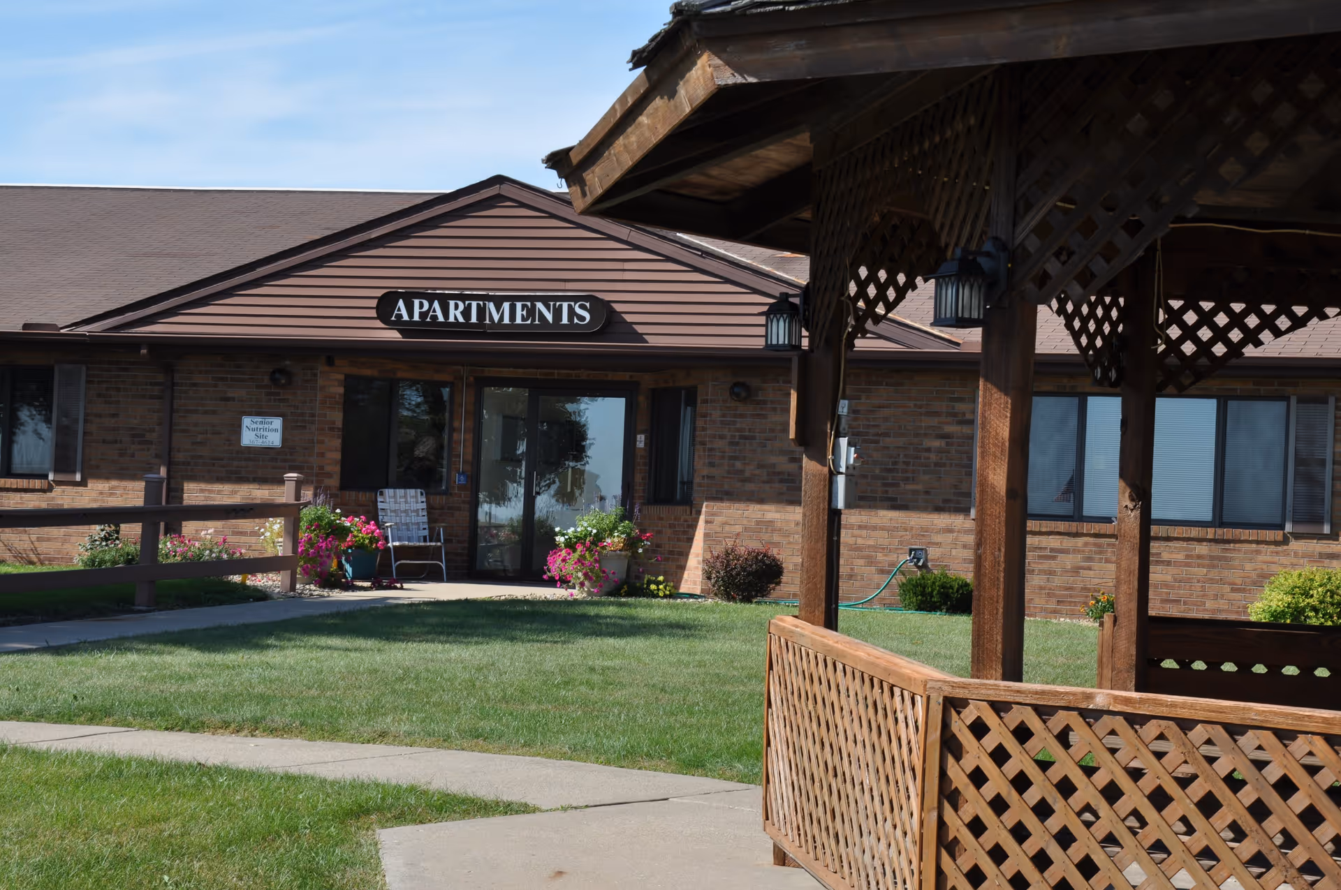 Exterior view of a brick building with a brown roof labeled 'APARTMENTS'. There is a well-maintained lawn with a concrete walkway and a wooden gazebo with lattice design in the foreground. Flower pots with colorful flowers are placed near the entrance.