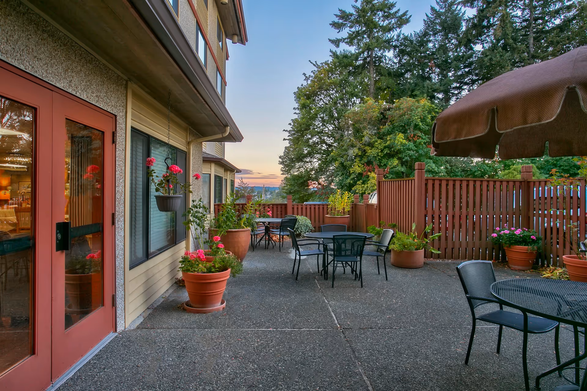Outdoor patio with metal tables and chairs, potted plants, and a wooden fence next to a senior living building at sunset.