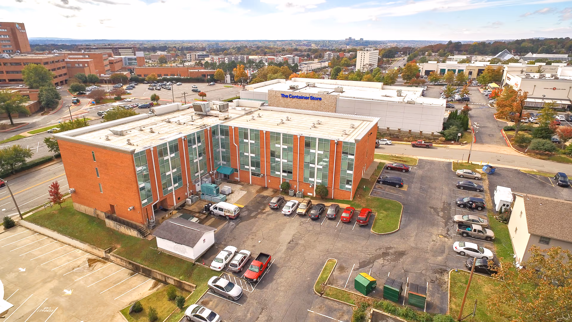 Aerial view of a three-story brick nursing facility with a parking lot, surrounding roads, and nearby commercial buildings.
