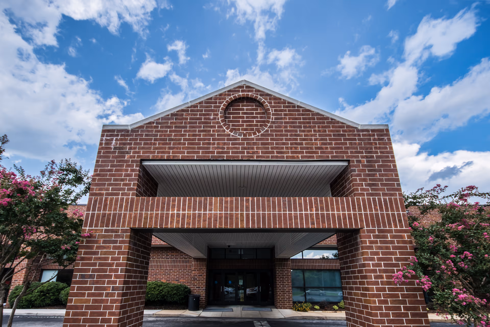 Front entrance of a red brick building with a covered portico, flowering shrubs, and a blue sky above.