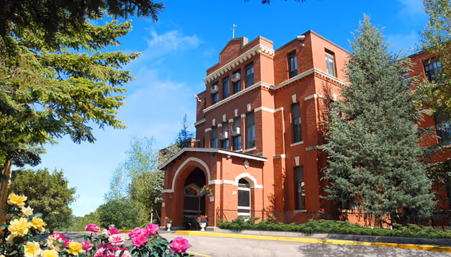 Red brick multi-story senior care building with an arched main entrance, flowering shrubs and tall trees under a blue sky.