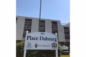 A white sign reading 'Place Dubourg' stands in front of a multi-story residential building with balconies under a clear blue sky.