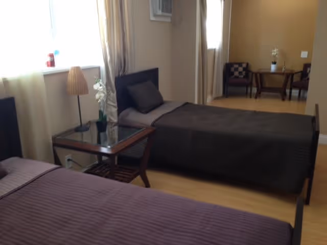 A bedroom with two single beds covered in dark bedspreads. Between the beds is a wooden side table with a glass top, a lamp, and a small potted plant. The room has light-colored walls and curtains, with a window letting in natural light. In the background, there is a small seating area with two chairs and a round table, also decorated with a small plant.