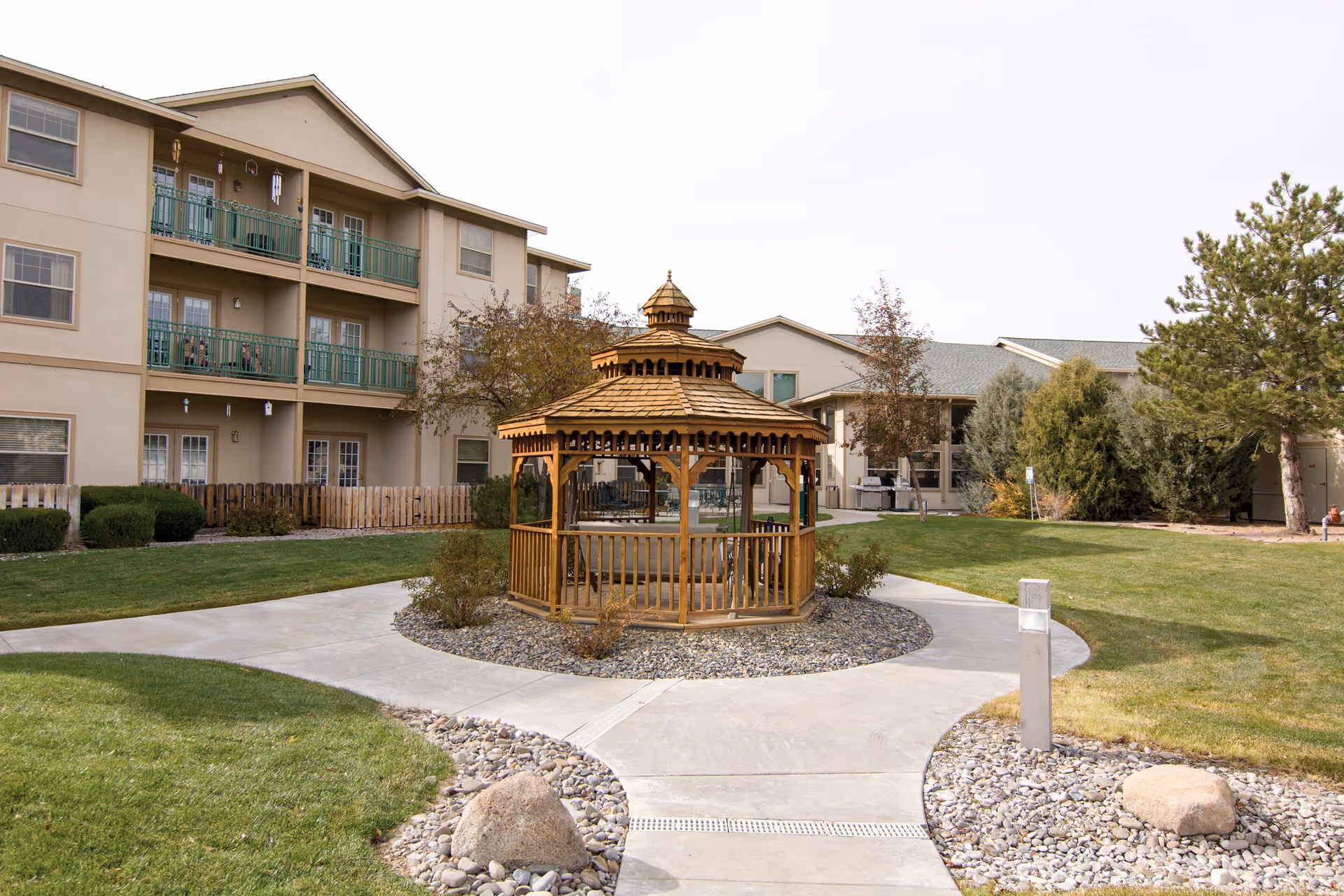 Outdoor area of The Chateau at Gardnerville featuring a wooden gazebo surrounded by a circular concrete walkway, green grass, bushes, and trees, with a multi-story residential building in the background.