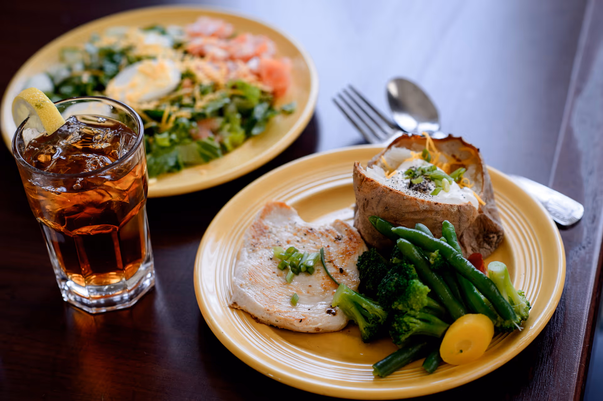 A meal consisting of a baked potato topped with sour cream and chives, steamed green beans, broccoli, and yellow squash, and a piece of grilled chicken garnished with chopped green onions on a yellow plate. Next to it is a glass of iced tea with a lemon wedge. In the background, there is a second yellow plate with a salad containing lettuce, sliced boiled eggs, shredded cheese, and tomato slices.