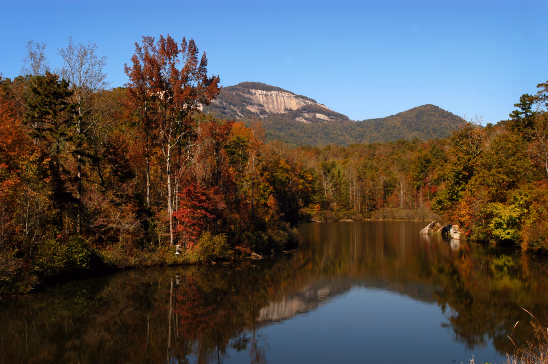 A serene outdoor scene featuring a calm lake reflecting the surrounding trees with autumn foliage and distant mountains under a clear blue sky.