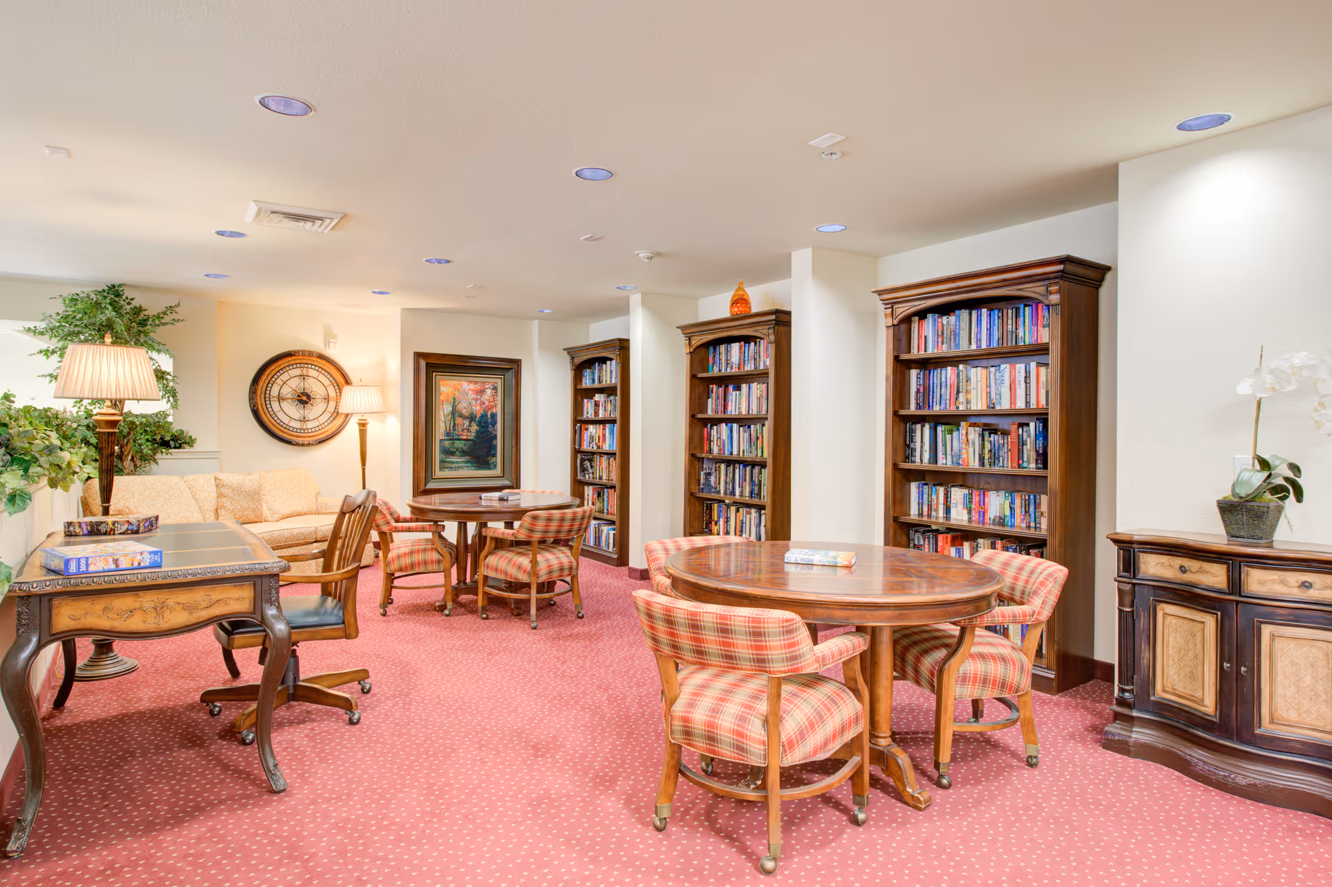 Bright senior living common room with round wooden tables and plaid chairs, bookshelves, a sofa, and lamps.