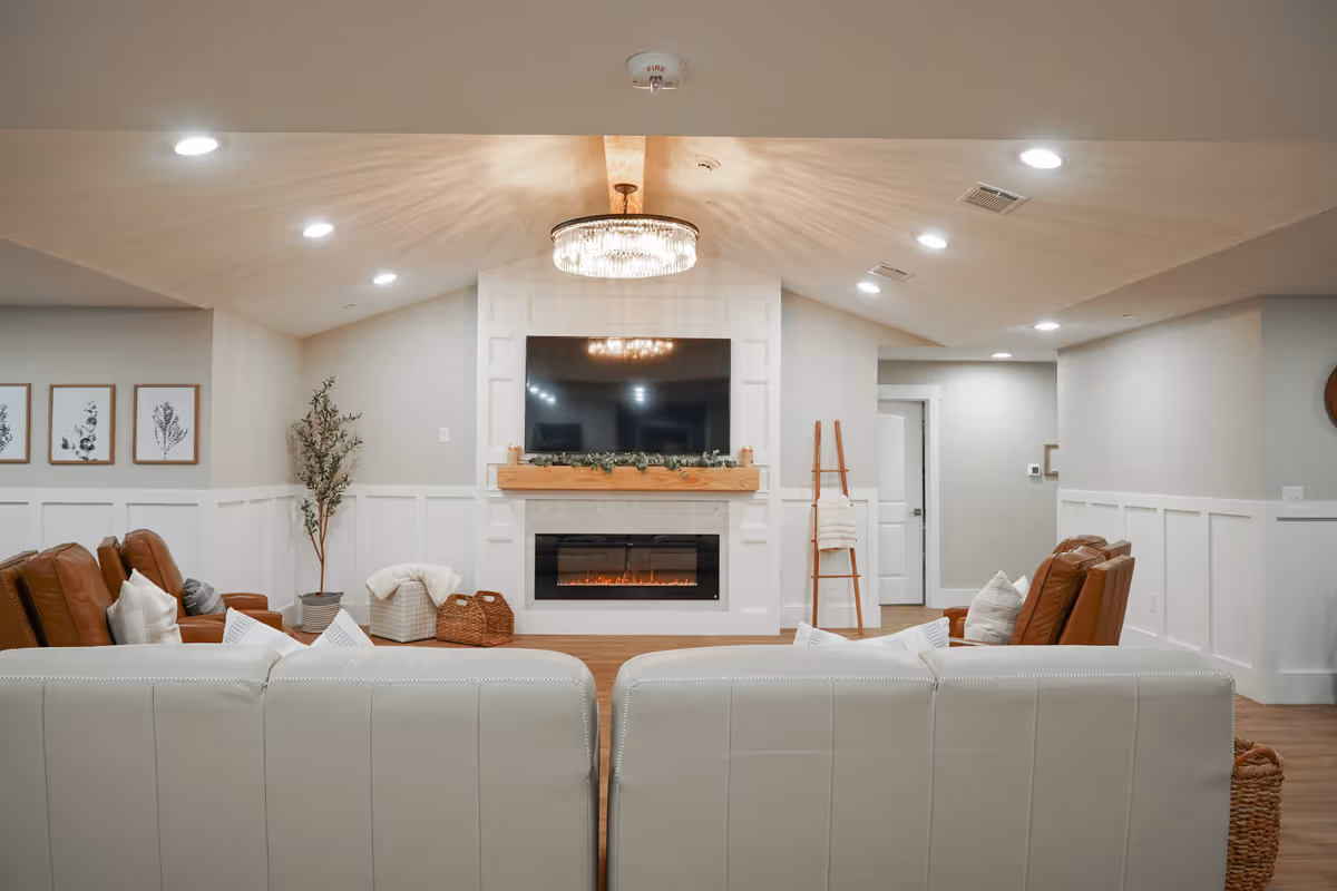 A cozy common living room with sofas and leather armchairs facing a wall-mounted TV above a fireplace beneath a decorative chandelier.