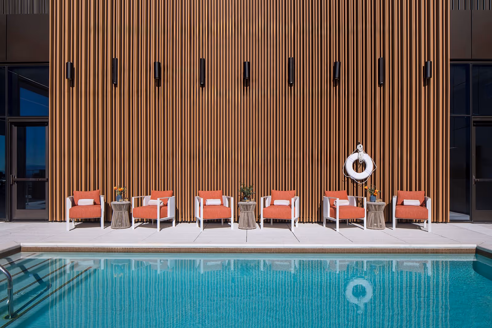 Outdoor swimming pool with clear blue water in front of a modern building wall featuring vertical wooden slats. Six orange cushioned chairs with white frames are arranged in a row along the poolside, each with a white towel on the seat. Three small round tables with flower vases are placed between the chairs. A white life preserver hangs on the wall above one of the tables.