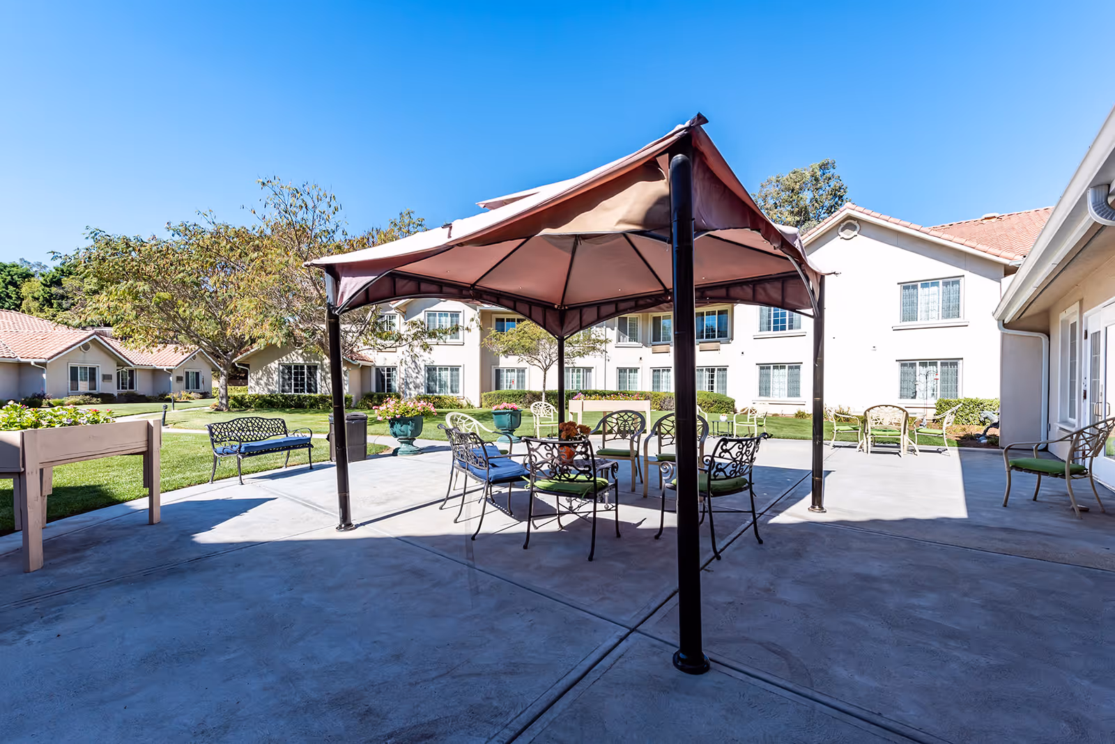 Outdoor patio area at The Palms At Bonaventure Assisted Living & Memory Care featuring a gazebo with metal chairs and tables, surrounded by green lawns, trees, and residential buildings under a clear blue sky.