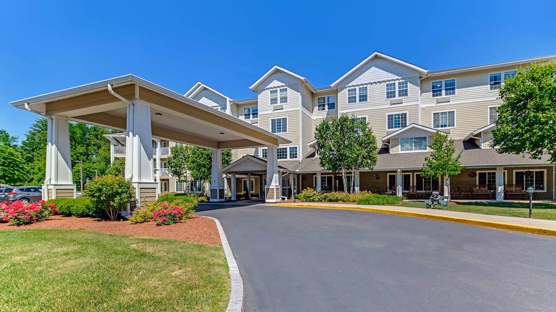 Exterior view of Holiday Birch Heights senior living facility on a sunny day, showing a multi-story beige building with white trim, a covered entrance driveway, landscaped greenery, and a clear blue sky.