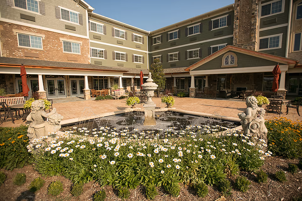 Outdoor courtyard area of The Bridge at Ooltewah featuring a central water fountain surrounded by white daisies and greenery, with stone cherub statues on either side. The courtyard is paved with brick and has seating areas with tables, chairs, and umbrellas. The multi-story building with green siding and brick accents surrounds the courtyard.
