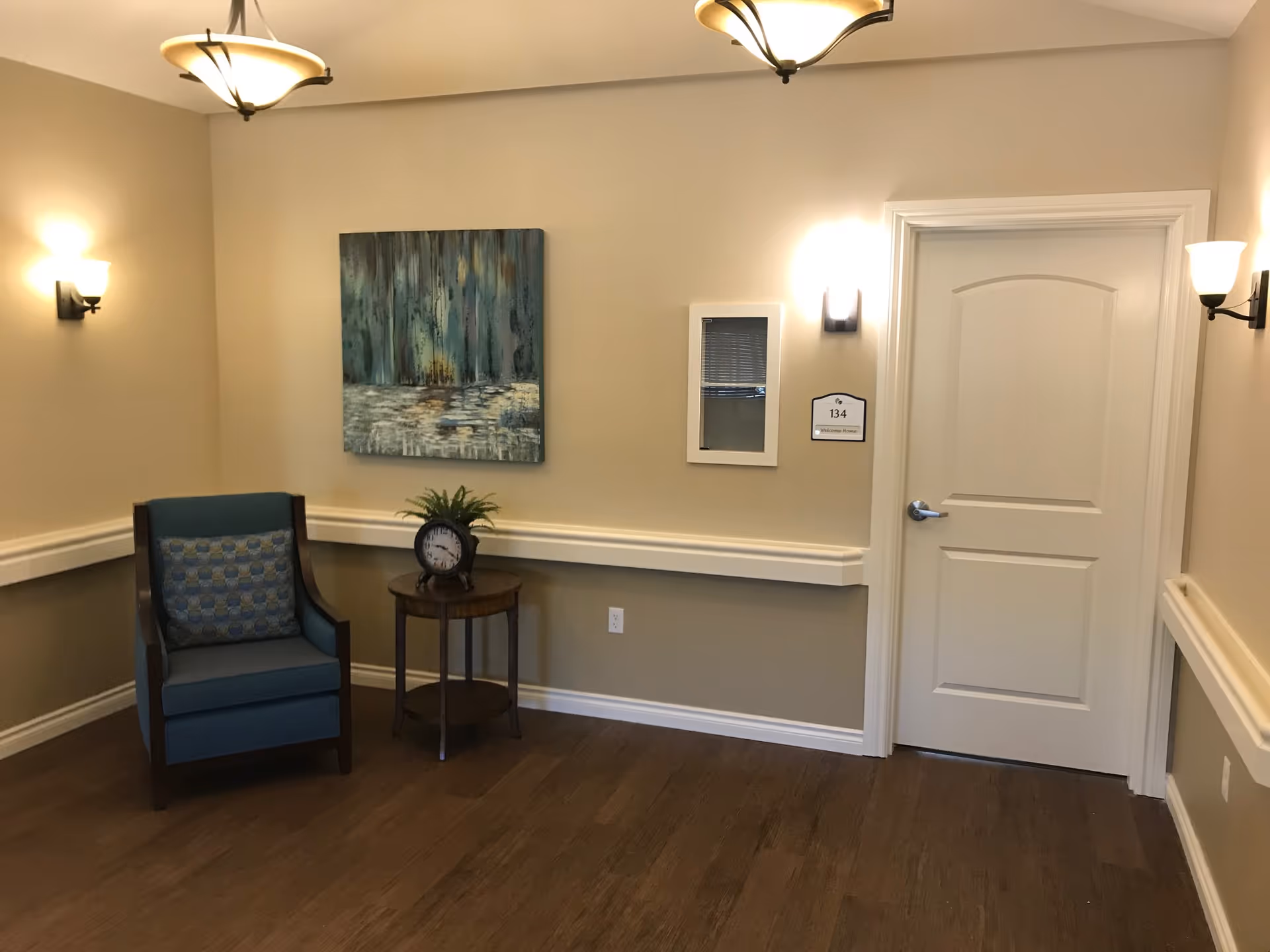 Small seating area with a blue armchair, round side table holding a clock and plant, wall art, and a closed door in a softly lit interior.