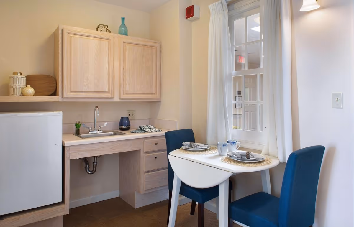 Small kitchenette and dining nook with a sink, light wood cabinets, mini fridge, and a two-seat table by a window.
