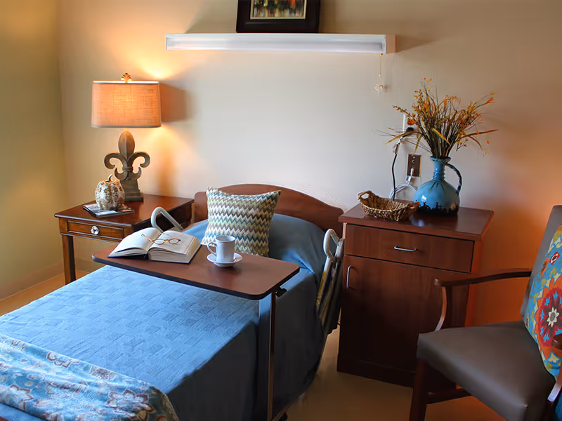 A tidy patient bedroom with a made bed covered in a blue blanket, an overbed table holding a cup and open book, bedside tables with a lamp and vase, and a chair.
