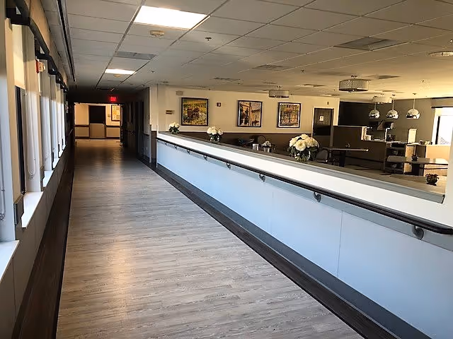 A long, well-lit hallway with wooden flooring and handrails on the right side. The hallway leads to a dining area with tables, chairs, and decorative flower arrangements. The walls have framed artwork, and there are ceiling lights throughout the space.
