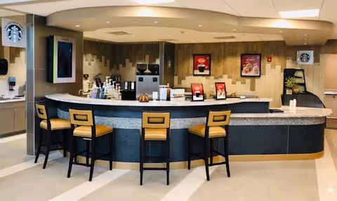 A modern coffee bar area with a curved counter and four high chairs with yellow cushions. Behind the counter are coffee machines, syrup bottles, and promotional signs. The walls have a textured design with framed coffee-themed posters and two Starbucks logos visible on the left and right sides.