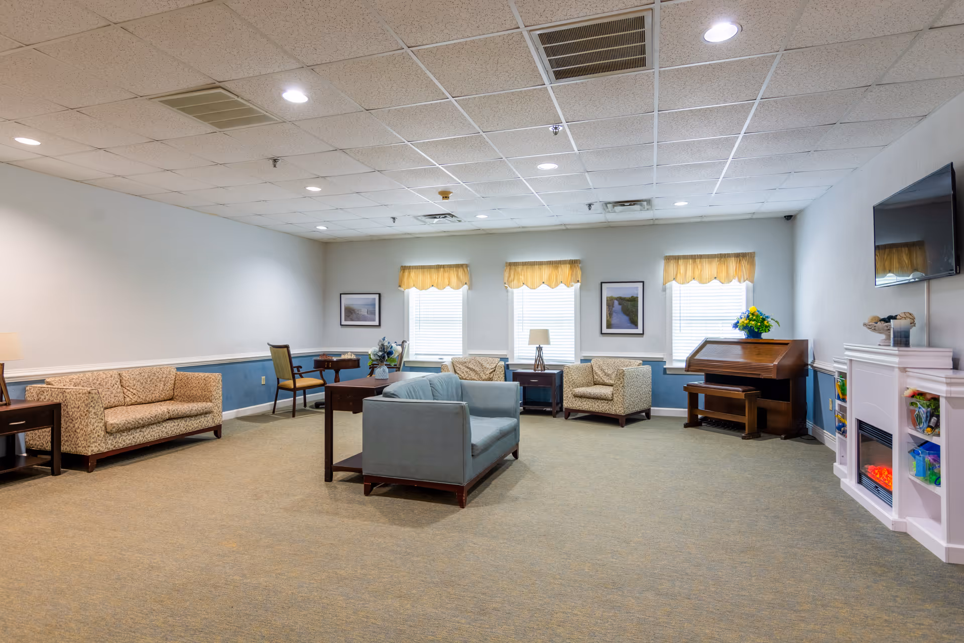 A spacious living room area in a senior care facility with multiple seating options including a blue loveseat, patterned armchairs, and a sofa. The room features three windows with yellow valances, framed pictures on the walls, a wooden piano with a bench, a flat-screen TV mounted on the wall, and a white electric fireplace with shelves containing toys and decorations.