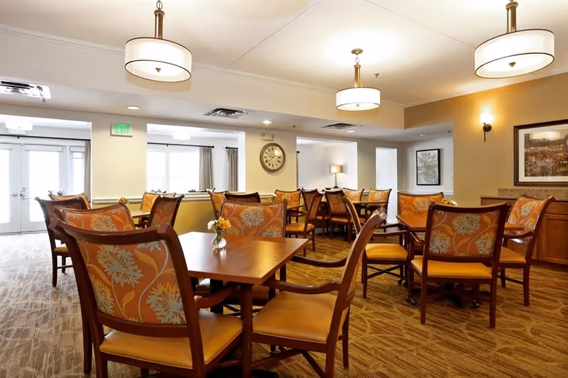 A well-lit dining room with multiple wooden tables and chairs featuring orange floral upholstery. The room has beige walls, carpeted flooring, and ceiling lights. There are framed pictures on the walls and a clock hanging in the background near large windows with curtains.