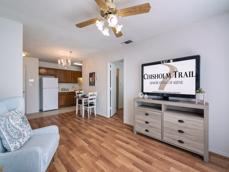 Interior view of a senior living apartment featuring a light blue armchair with a patterned pillow, a wooden floor, a small dining table with two chairs, a kitchen area with wooden cabinets and a white refrigerator, and a TV on a wooden stand displaying the Chisholm Trail Senior Living logo.