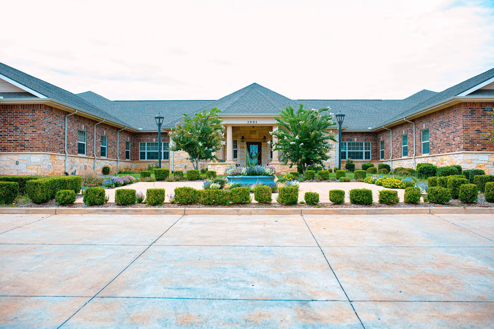Front entrance of a single-story brick building with a central portico, fountain, and landscaped shrubs.