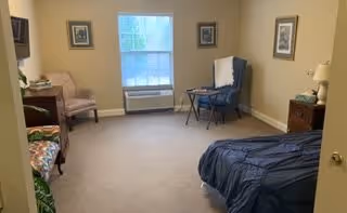 A simple bedroom with beige walls and carpeted floor. The room contains a bed with a dark blue bedspread on the right side, a wooden nightstand with a lamp, a blue reclining chair with a white blanket draped over it, a small table, and a beige armchair near a window with closed blinds. There are framed pictures hanging on the walls.