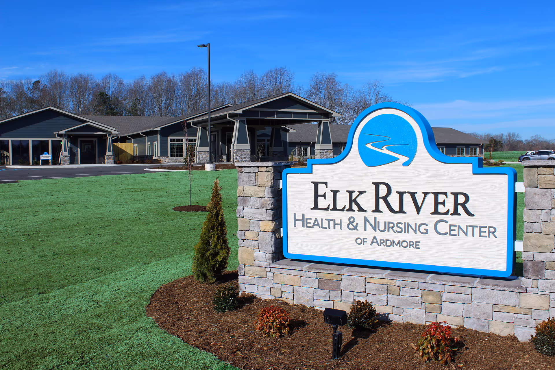 Exterior view of Elk River Health & Nursing Center of Ardmore with a large sign in the foreground and the building in the background under a clear blue sky.