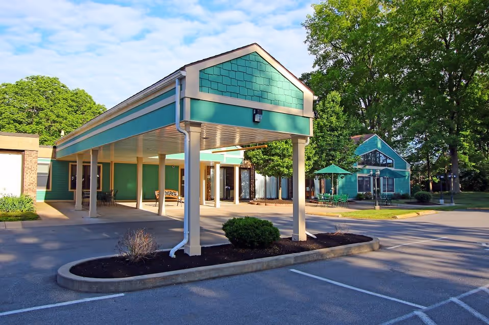 Exterior view of a senior living facility with a covered entrance supported by columns, green siding, and a small landscaped area with bushes. In the background, there is a building with large windows and outdoor seating with green umbrellas surrounded by trees.