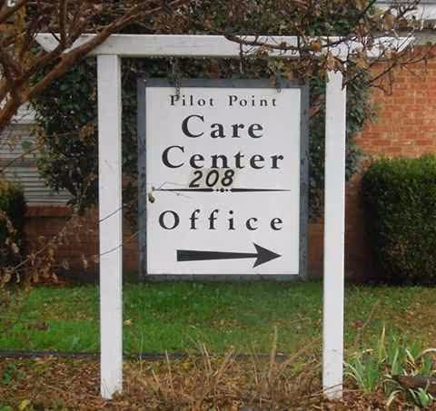 White framed sign reading Pilot Point Care Center Office with arrow, in front of brick building.