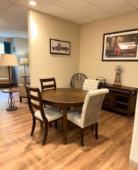 A cozy interior space with a round wooden dining table surrounded by four chairs, two with wooden backs and two upholstered. The room has wood flooring and beige walls. On the wall, there is a framed picture of a red barn and a decorative windmill on a wooden sideboard. A 'welcome' sign is also mounted on the wall. In the background, there is a sitting area with a lamp and armchair near a window with blue curtains.