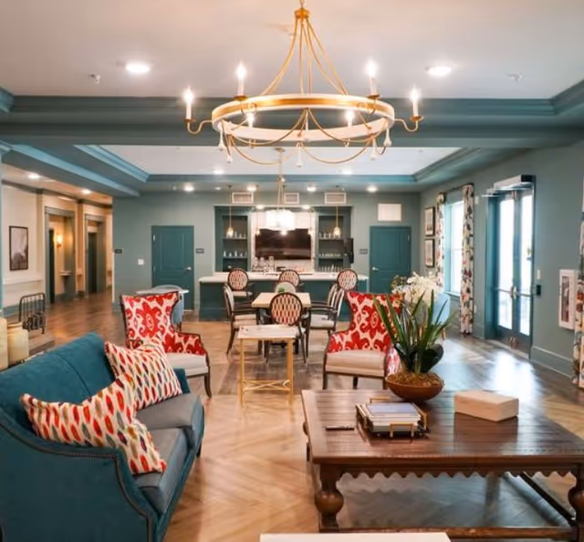 A spacious and well-lit common area with teal walls and ceiling accents, featuring a large chandelier. The room has a teal sofa with patterned pillows, two red and white patterned armchairs, a wooden coffee table with books and a plant, and a dining area with several chairs and tables. There are large windows with patterned curtains and double glass doors letting in natural light.