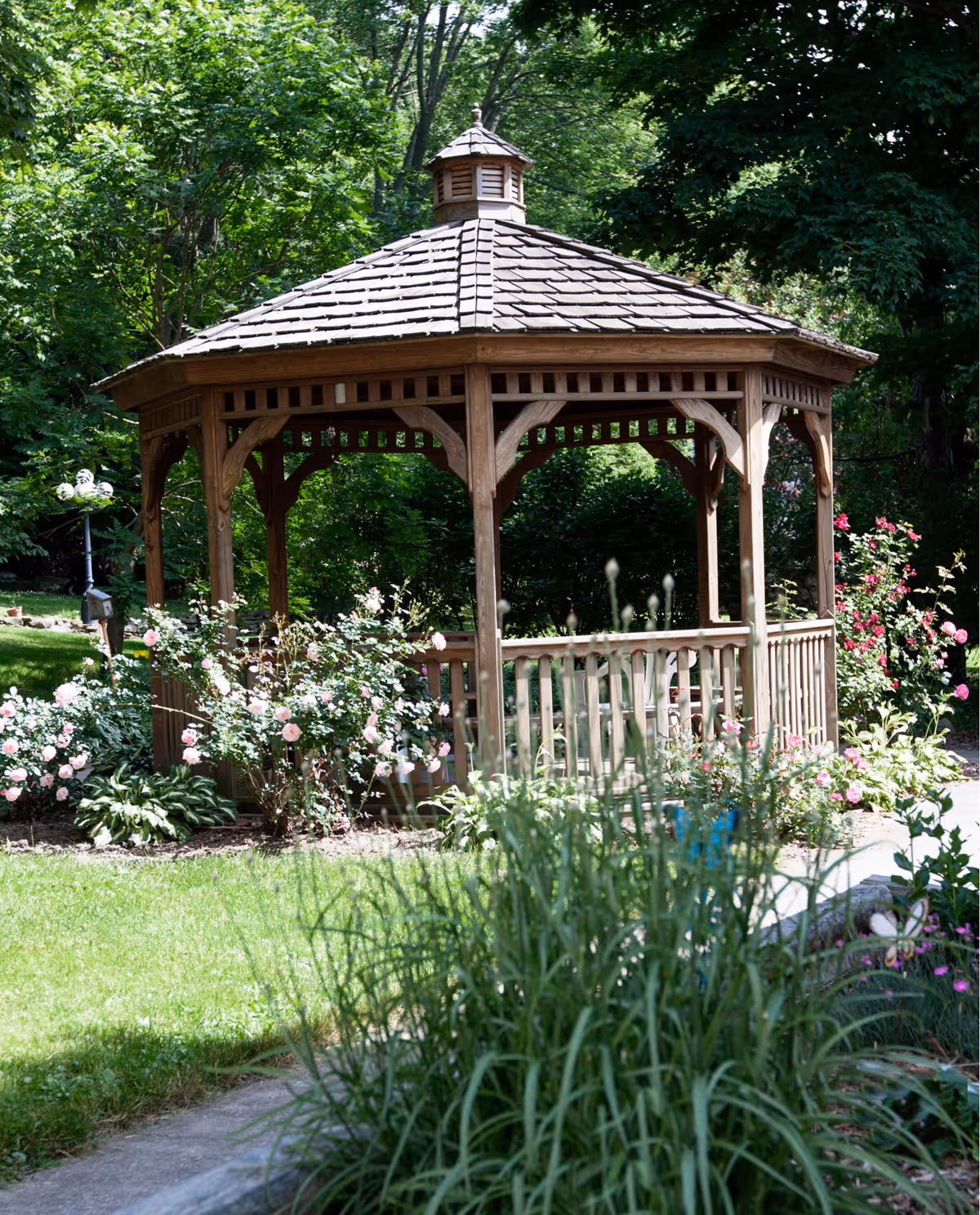 A wooden gazebo with a shingled roof surrounded by green grass, flowering bushes, and trees in a garden setting.