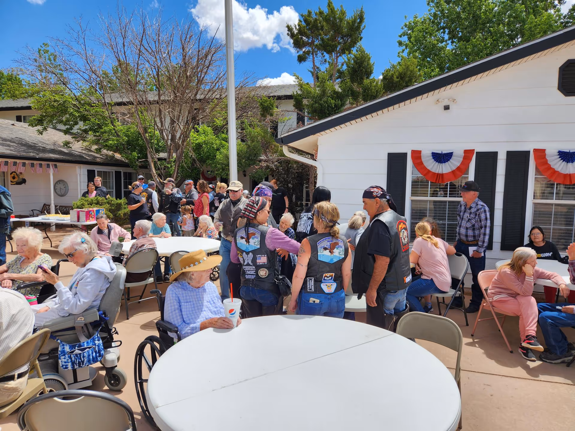 Outdoor gathering at Carriage House On West Garden Lane with elderly residents and visitors seated and standing around tables on a sunny day. The building is decorated with red, white, and blue bunting, and people are engaged in conversation and activities.