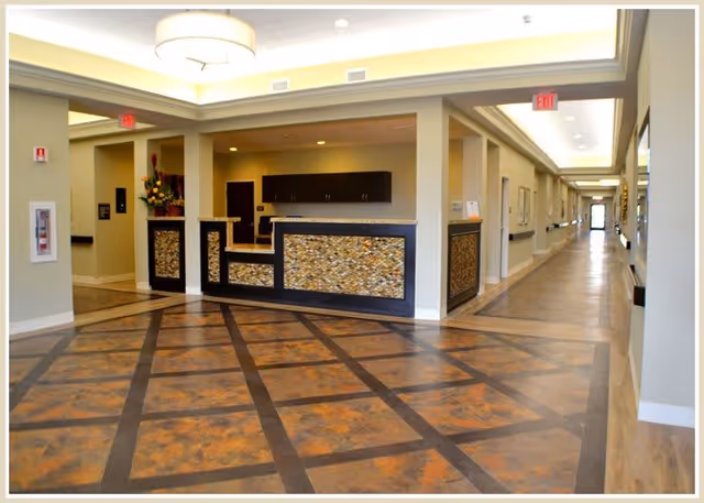 Reception area with a decorative front desk featuring mosaic panels, a patterned floor with brown and orange tones, beige walls, ceiling lights, and a long hallway extending to the right with handrails along the walls.