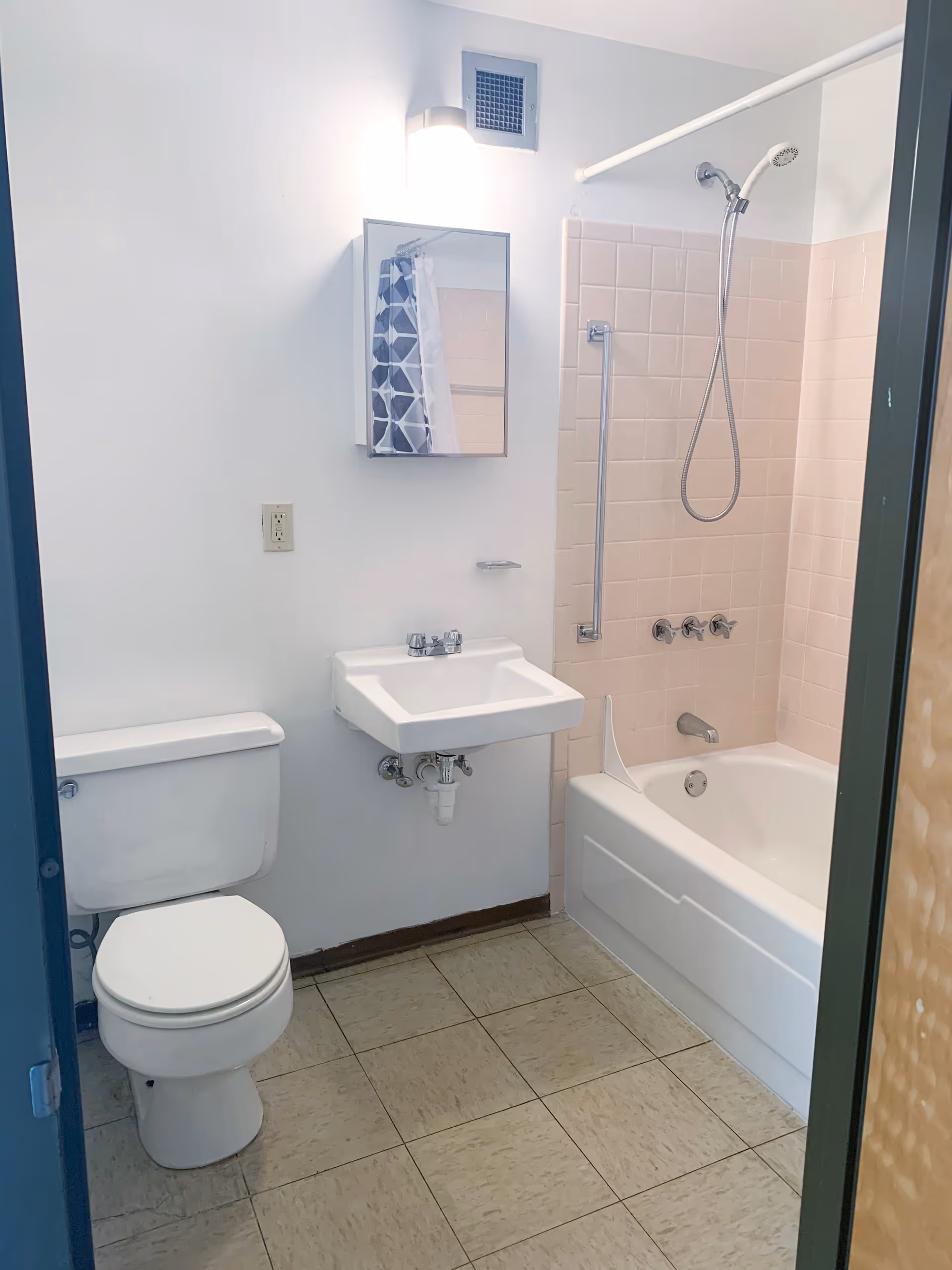 A small bathroom with a white toilet, a wall-mounted sink, a mirror cabinet above the sink, and a bathtub with a showerhead and pink tiled walls. The floor is covered with beige tiles.