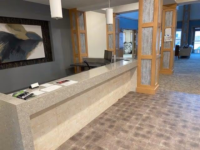 Reception desk area with a long countertop, a computer monitor, and some papers on the counter. The area features wooden framed glass panels and a carpeted floor extending into a hallway with seating visible in the distance. A large abstract painting is mounted on the wall behind the desk.