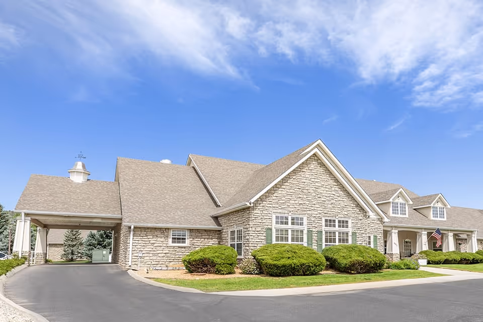 Exterior view of a single-story senior living facility building with stone facade, multiple windows, green shrubs, and a covered driveway under a blue sky with some clouds.
