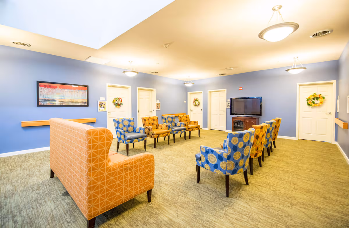 Well-lit communal lounge with patterned armchairs and an orange sofa arranged facing a wall-mounted TV.