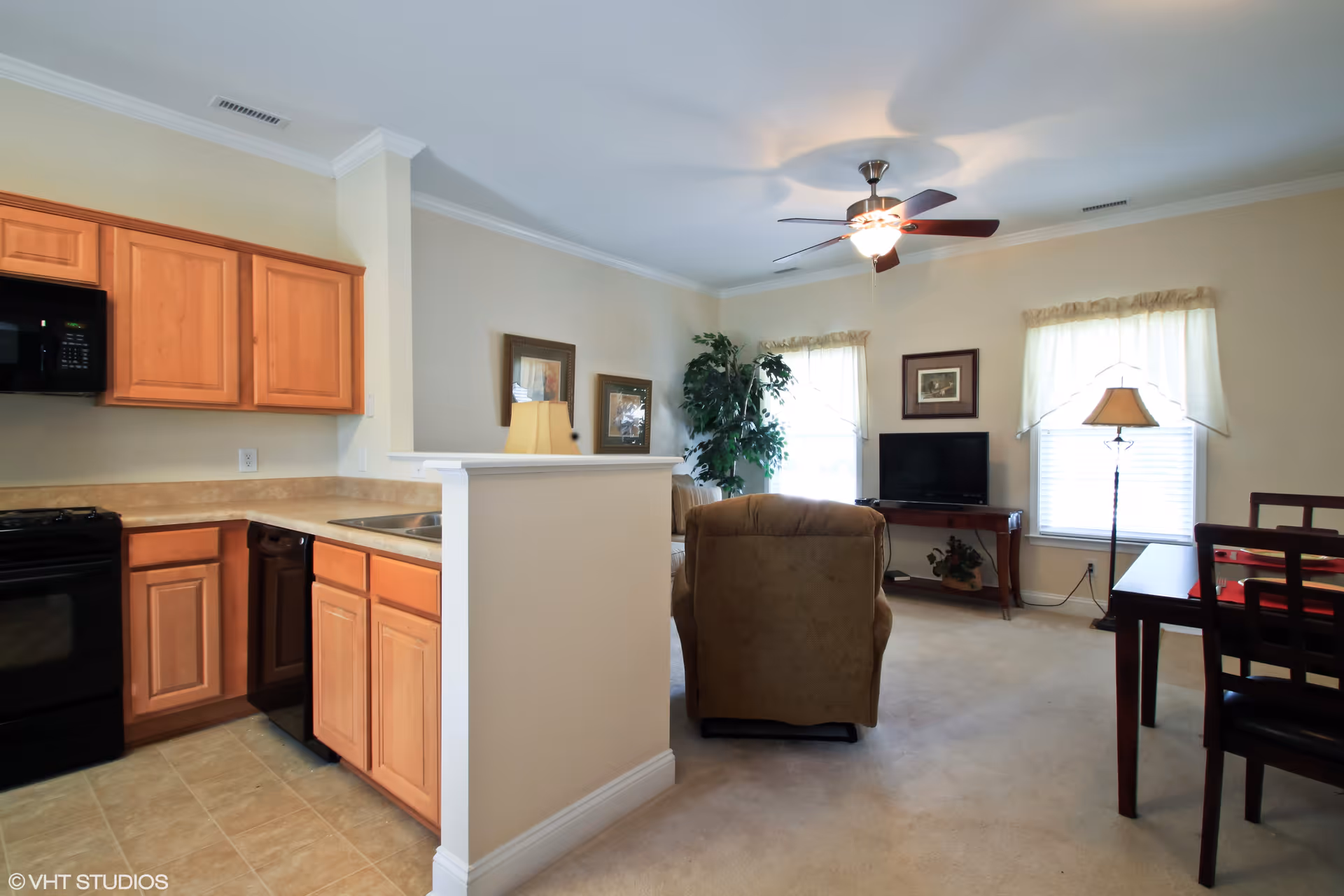 Open-plan living area with a small kitchen, recliner facing a TV, dining table, and ceiling fan.