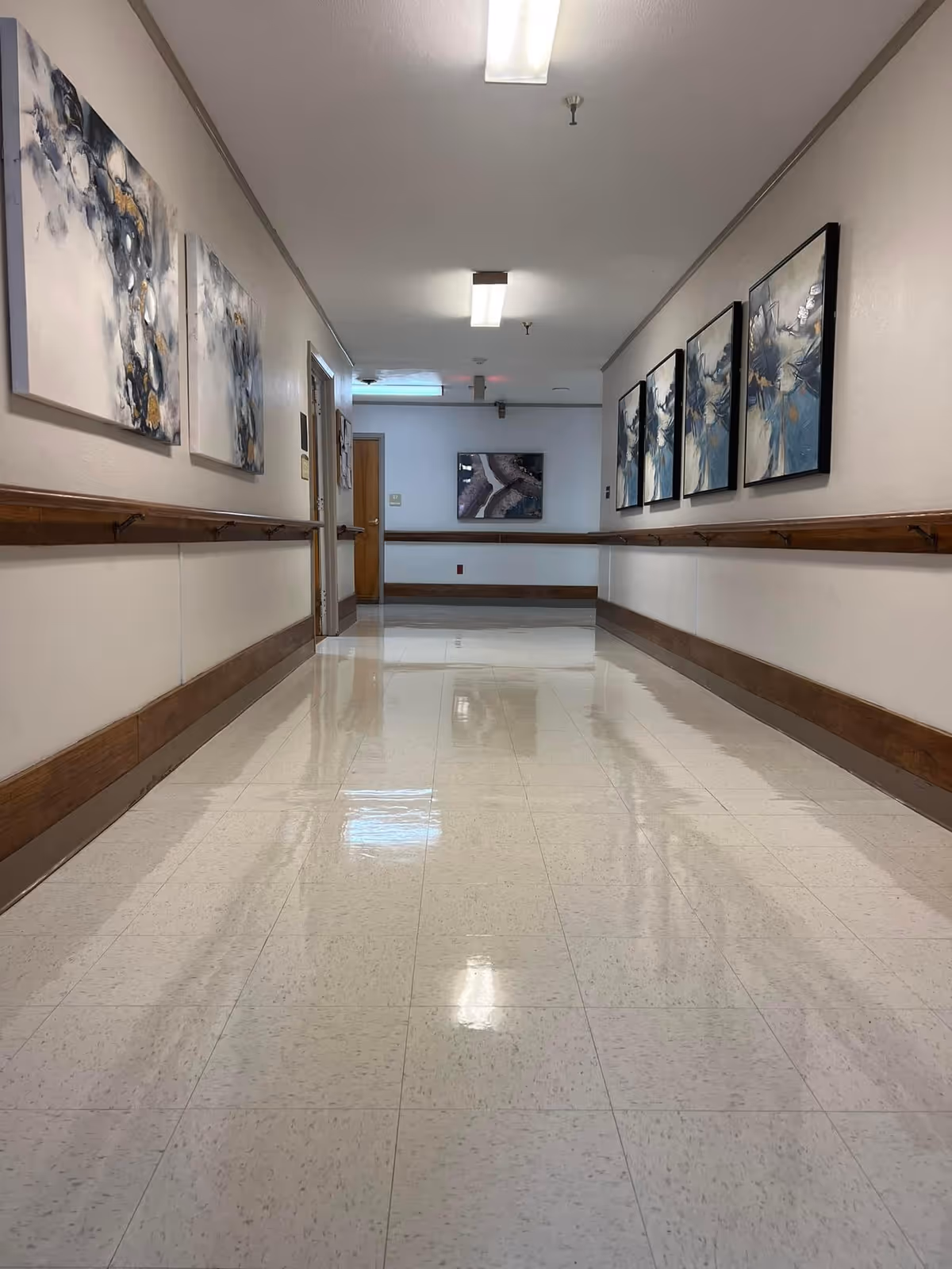 A clean, well-lit hallway in a nursing and rehabilitation facility with polished tile floors, handrails along both walls, and abstract artwork hanging on the walls.