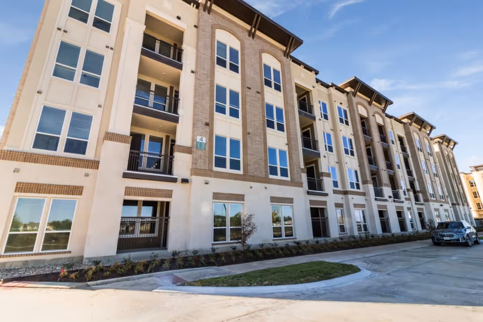 Exterior view of a multi-story residential building with large windows and balconies, a driveway with a parked car, and a clear blue sky.
