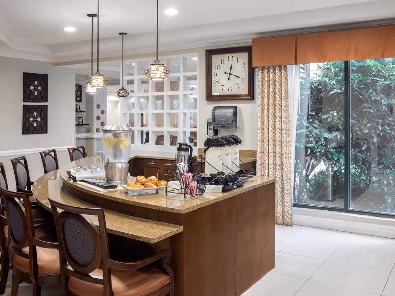 A well-lit dining area with a curved wooden counter and several chairs. On the counter, there is a large glass dispenser filled with water and lemon slices, a tray with pastries, coffee cups, and various condiments. Behind the counter, there is a wall clock, a paper towel dispenser, and a window with curtains showing greenery outside. The room has pendant lights hanging from the ceiling and decorative wall art.